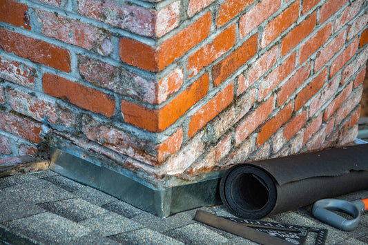New metal flashing around the base of a brick chimney on an asphalt shingle roof, with a roll of underlayment beside it