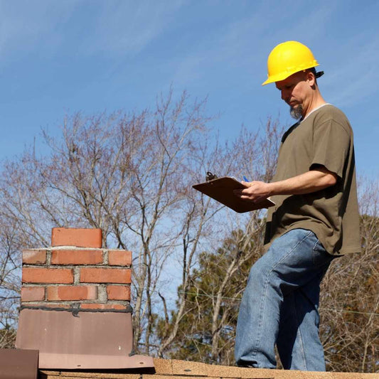 Roofer in a yellow hard hat with a clipboard inspecting a roof beside a brick chimney