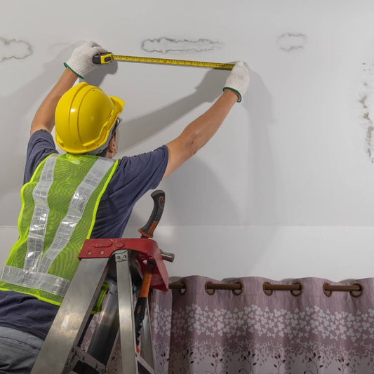 Worker in a hard hat on a ladder measuring water stains near the top of an interior wall