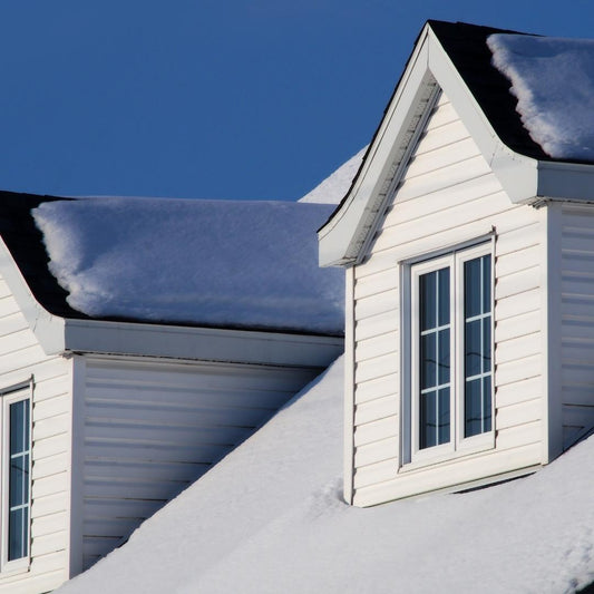 Snow covering the roof and dormer of a house with white siding under a clear blue sky