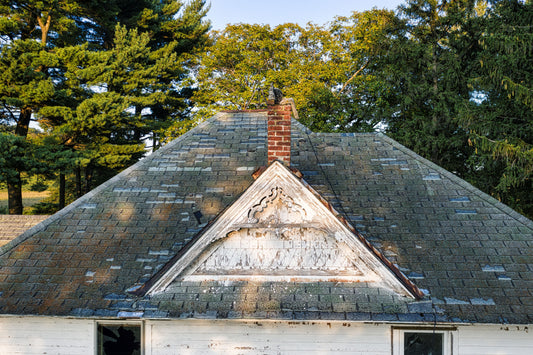 Older asphalt shingle roof with worn, missing shingles and moss around a brick chimney, with pine trees behind it