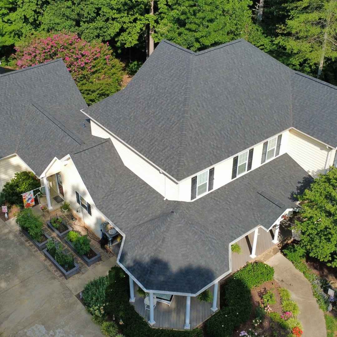 An aerial view of a multi-gabled house with dark gray asphalt or composite roofing, white siding, and a landscaped yard with trees and flower beds.