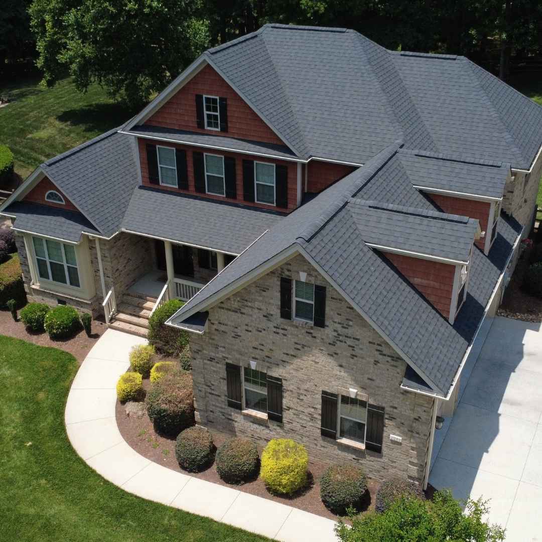 An aerial view of a two-story residential house with a dark gray shingled roof, red brick upper walls, and a stone lower facade, set in a landscaped yard with lawn, shrubs, and a curved walkway.